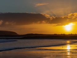 A serene sunset over a coastal landscape with a silhouette of a distant hill. The sun sets behind clouds, casting a golden glow on the sea and reflecting on the wet sand.