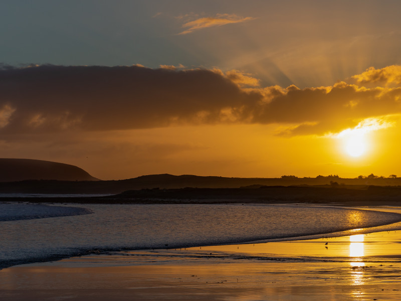 A serene sunset over a coastal landscape with a silhouette of a distant hill. The sun sets behind clouds, casting a golden glow on the sea and reflecting on the wet sand.