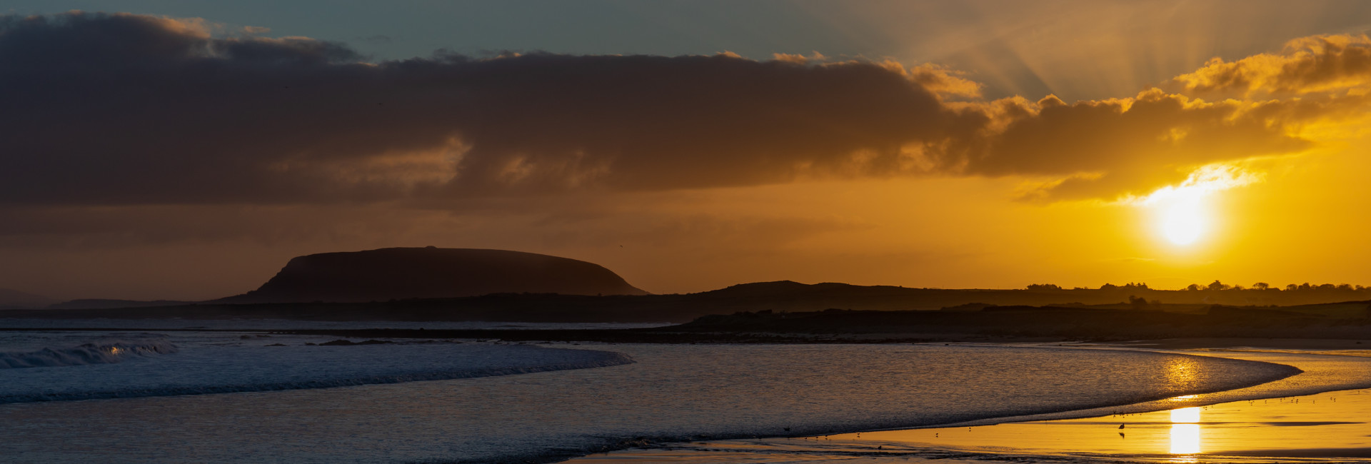 A serene sunset over a coastal landscape with a silhouette of a distant hill. The sun sets behind clouds, casting a golden glow on the sea and reflecting on the wet sand.