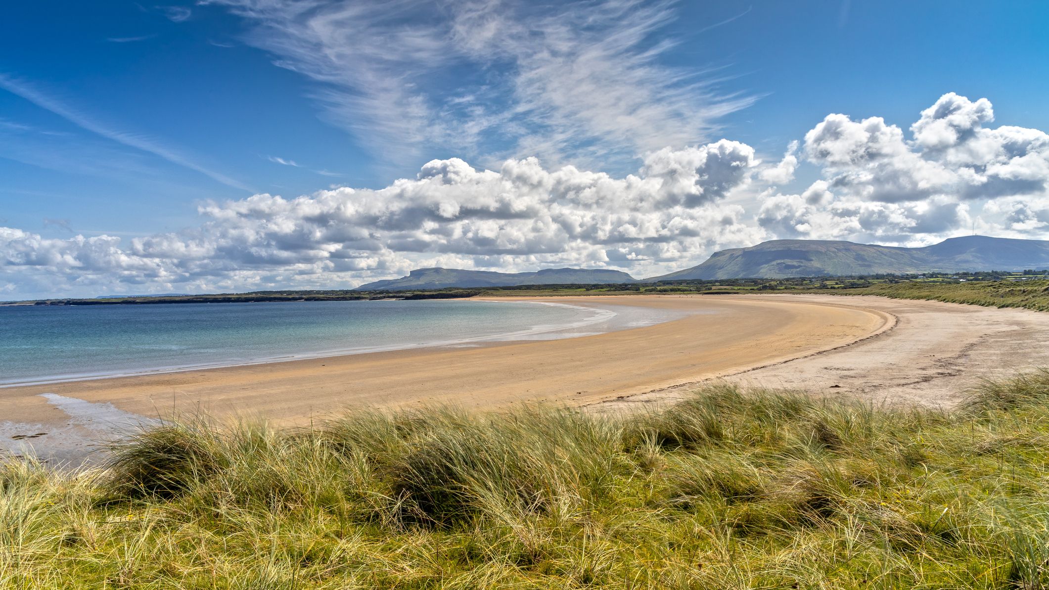 A picturesque beach with golden sands and calm, clear waters. Grassy dunes in the foreground lead to a wide, sweeping bay under a sky filled with fluffy white clouds.