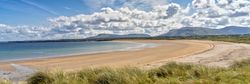 A picturesque beach with golden sands and calm, clear waters. Grassy dunes in the foreground lead to a wide, sweeping bay under a sky filled with fluffy white clouds.