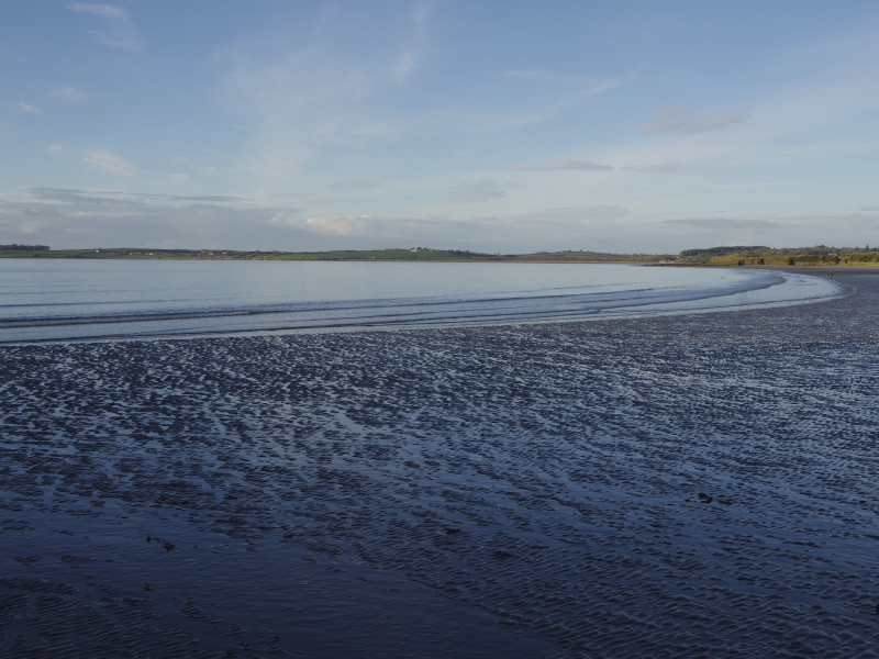 A beach at low tide, where rippled sand meets still, shallow water under a tranquil sky. Distant grassy hills line the horizon, adding depth to the expansive view.