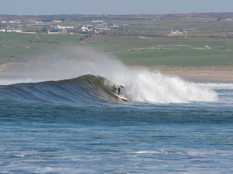 A surfer riding a breaking wave on a sunny day with a green coastal landscape and scattered houses in the background. The wave is curling, creating a spray of white water.
