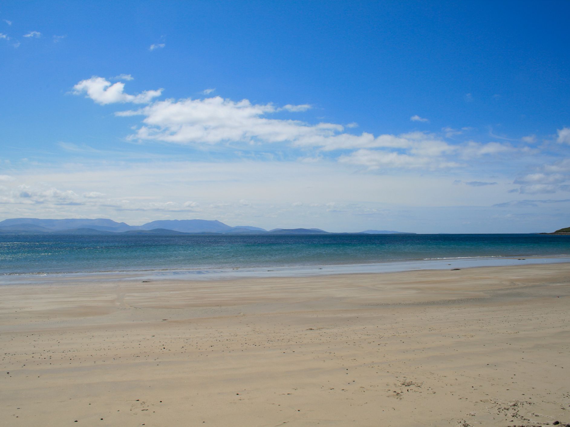 A serene beach with soft sand stretches toward calm, blue-green ocean waters under a bright blue sky with scattered clouds. Distant mountains line the horizon in the background.