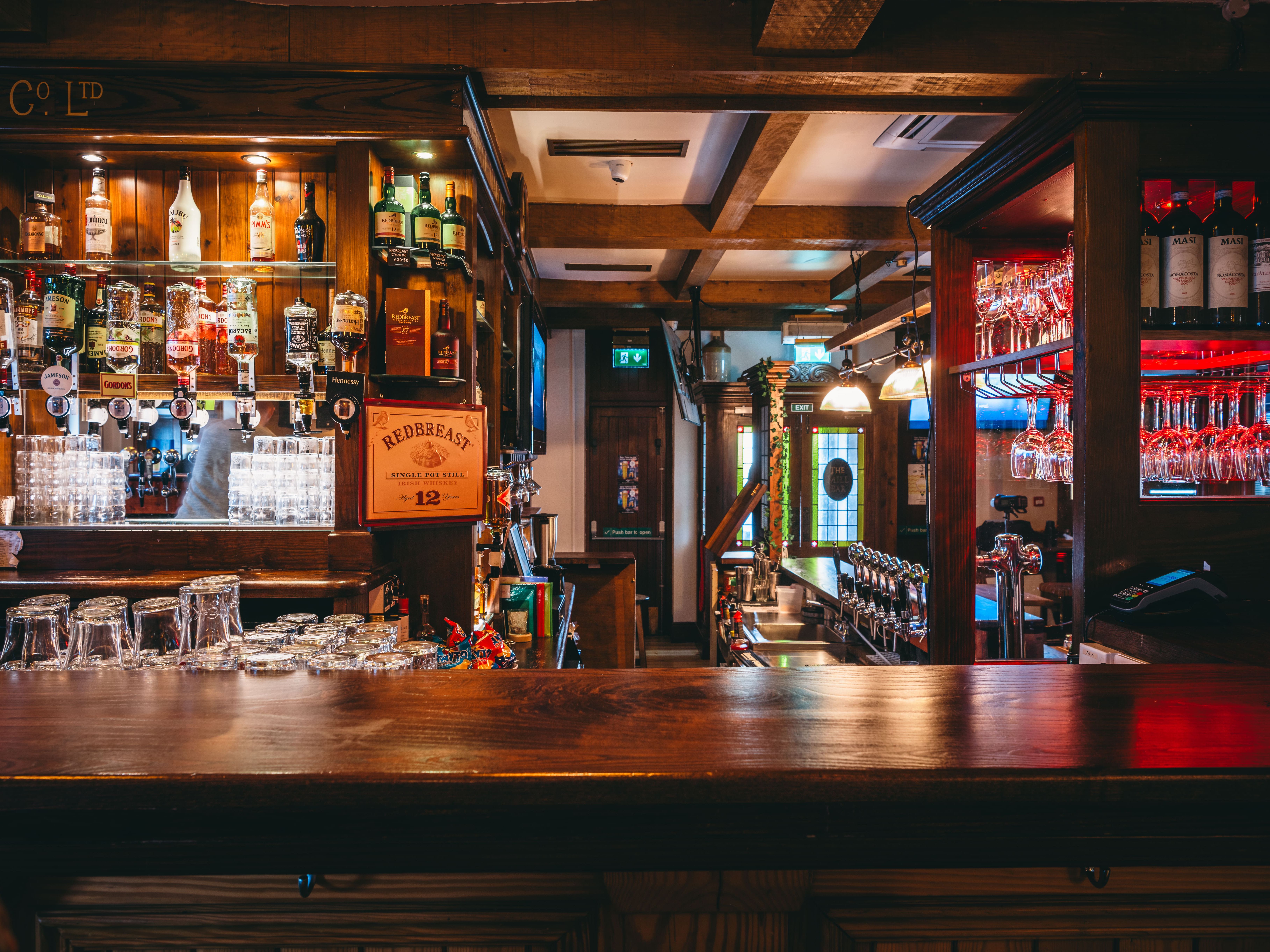 An empty bar with rows of liquor bottles on shelves, glassware neatly arranged, and taps lined up. Warm lighting highlights the wooden beams and classic pub decor.