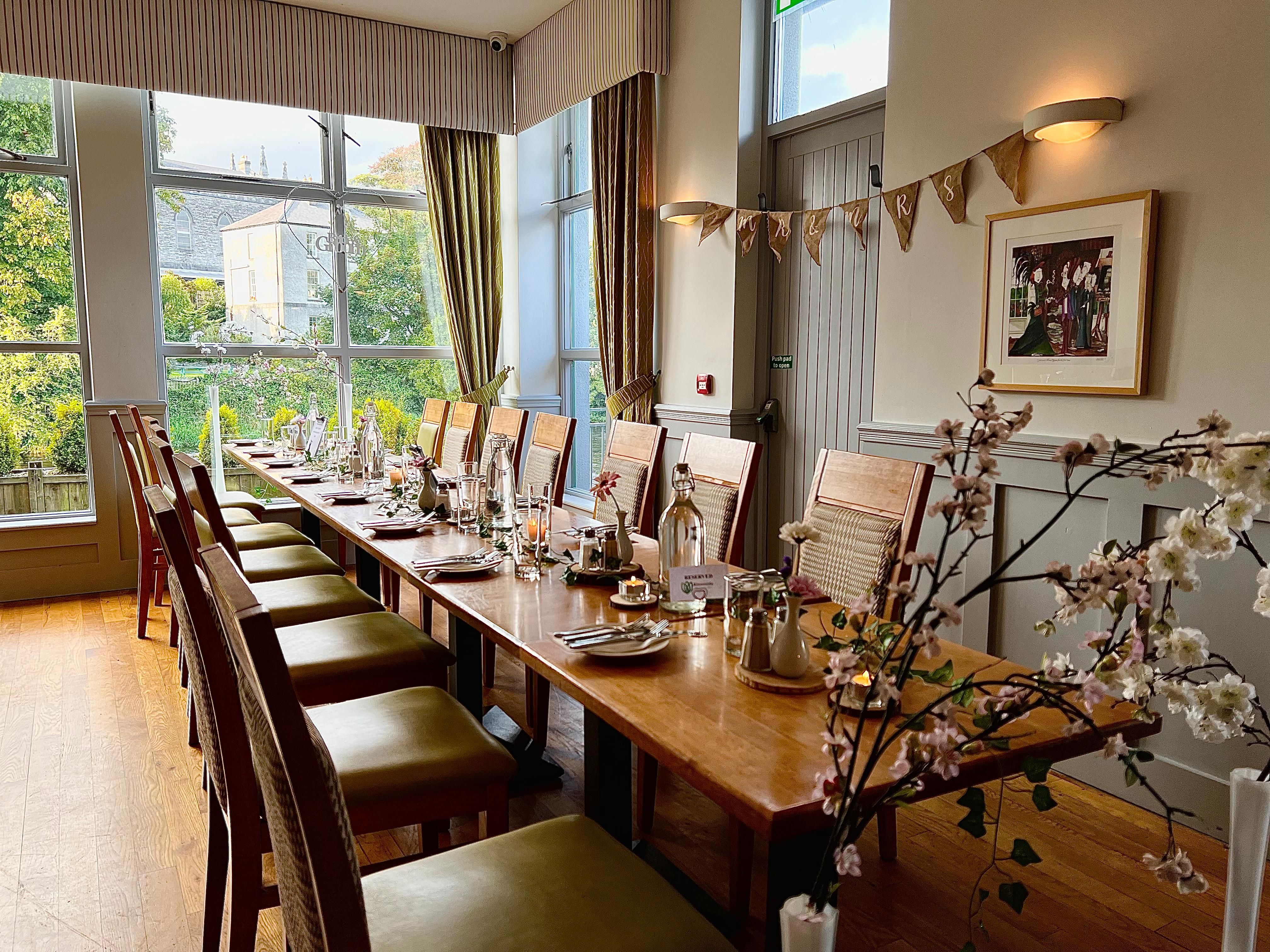 A long dining table is elegantly set with plates, glasses, and centerpieces in a bright room with large windows and a garden view. A
