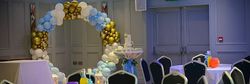 A decorated event room with round tables, black chairs, and colorful balloons. A balloon arch in white, gold, and blue sits in the background, along with a tall table and lanterns.