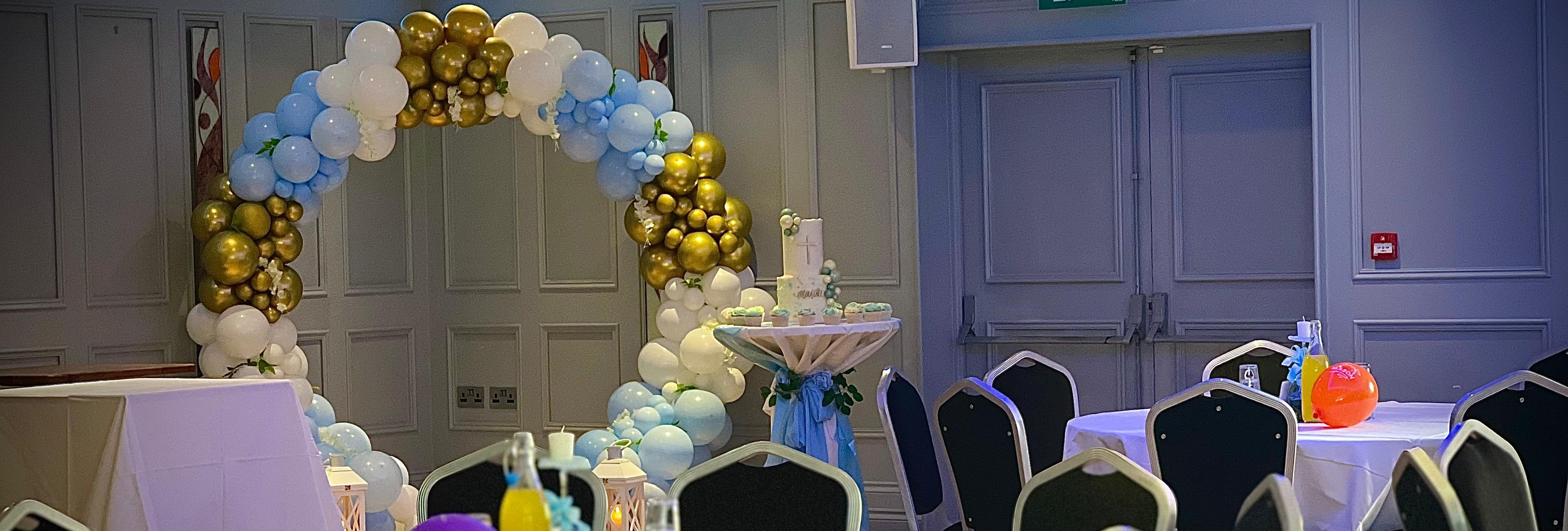 A decorated event room with round tables, black chairs, and colorful balloons. A balloon arch in white, gold, and blue sits in the background, along with a tall table and lanterns.