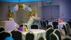 A decorated event room with round tables, black chairs, and colorful balloons. A balloon arch in white, gold, and blue sits in the background, along with a tall table and lanterns.