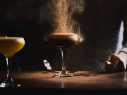 A bartender sprinkles a fine powder over a dark cocktail in a coupe glass, creating a cloud effect. Another yellow cocktail sits on the side, both on a dimly lit bar.