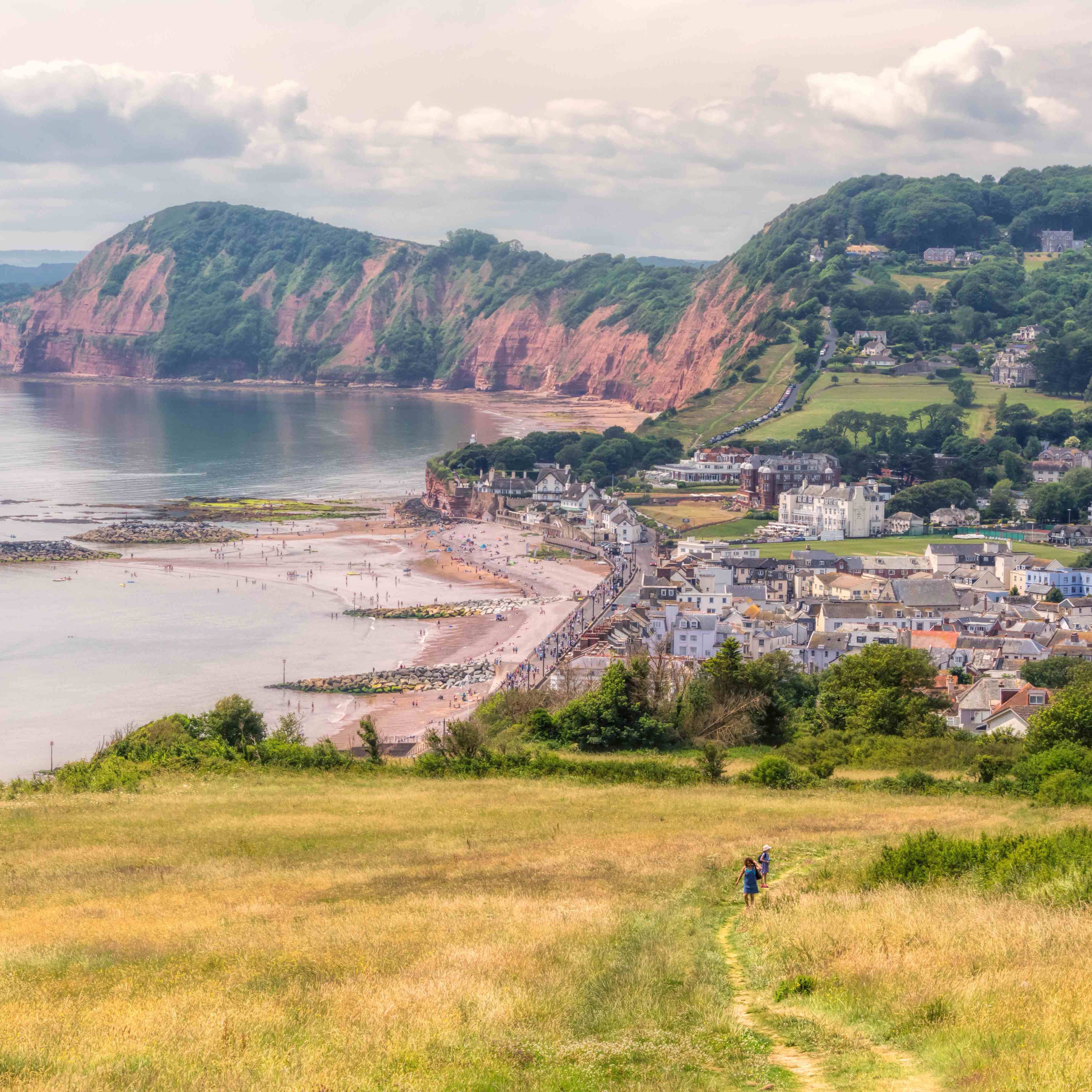 A scenic view of Sidmouth, Devon, featuring the town, sandy beach, and iconic red cliffs of the Jurassic Coast, captured from a grassy hilltop
