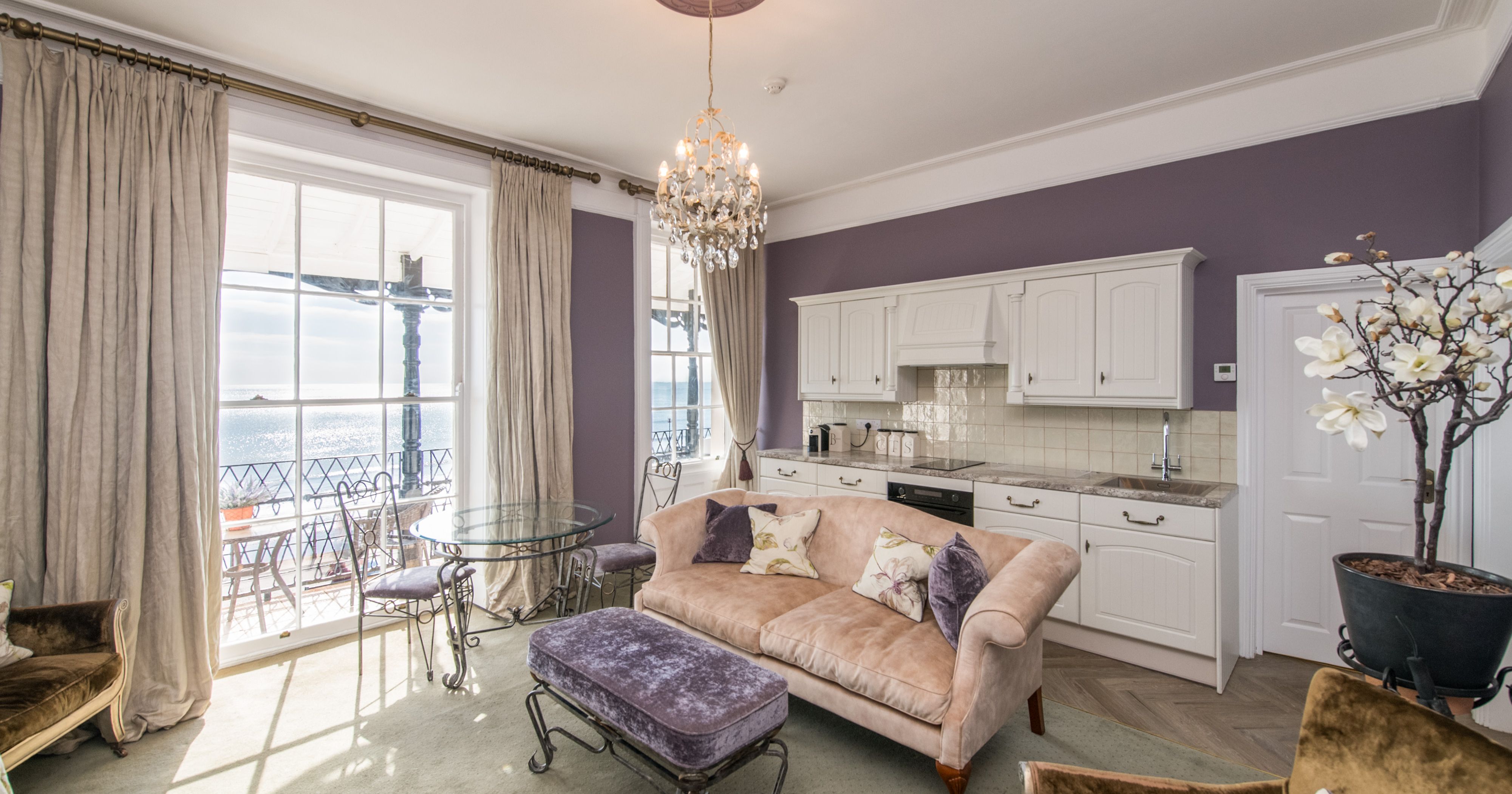Elegant living room and kitchen with purple walls, chandelier, and sea view through large windows.