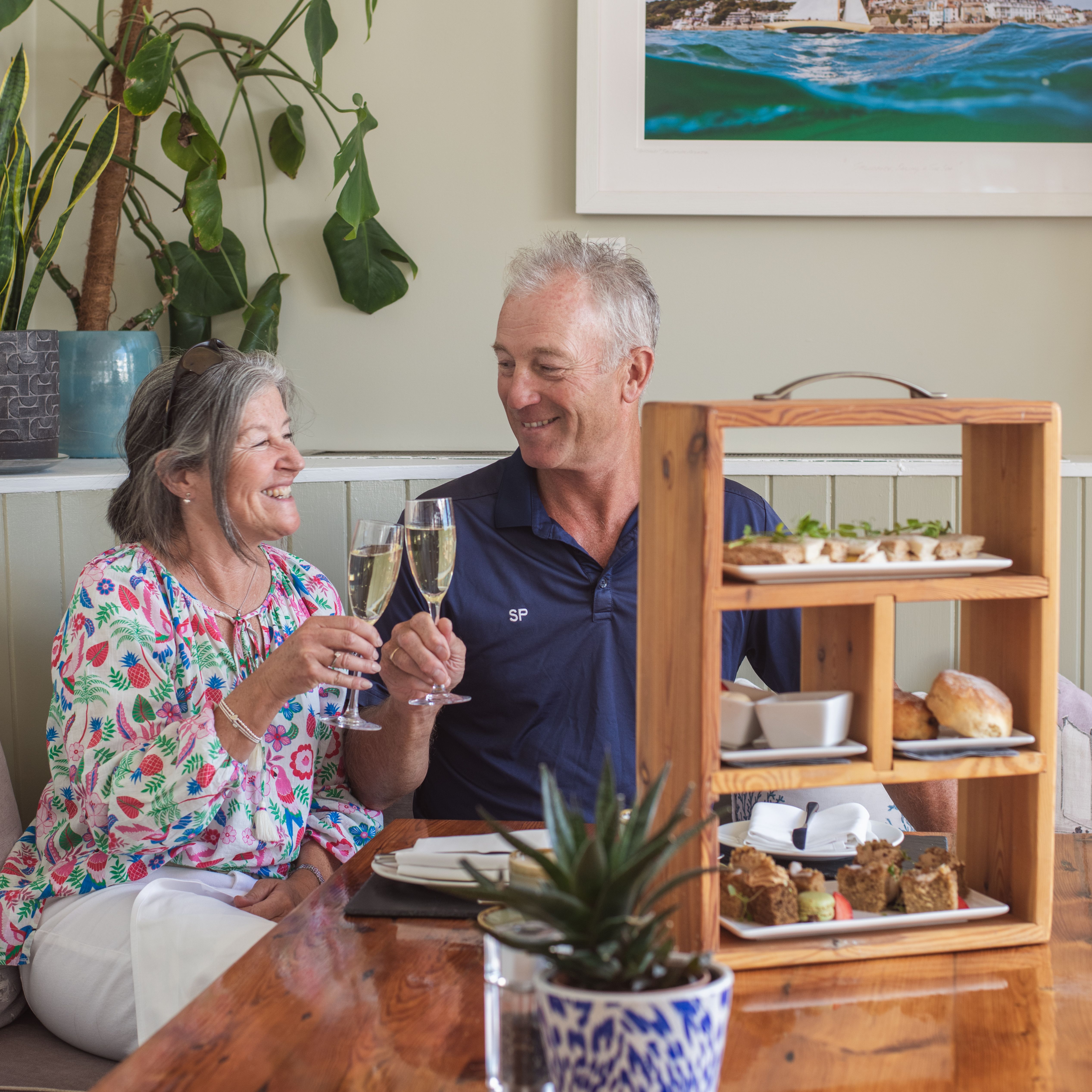 Older couple smiling and clinking champagne glasses while enjoying afternoon tea together at a cozy restaurant.