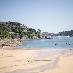 A sandy beach with people walking and relaxing, a calm blue sea with boats, and green hills with houses in the background.