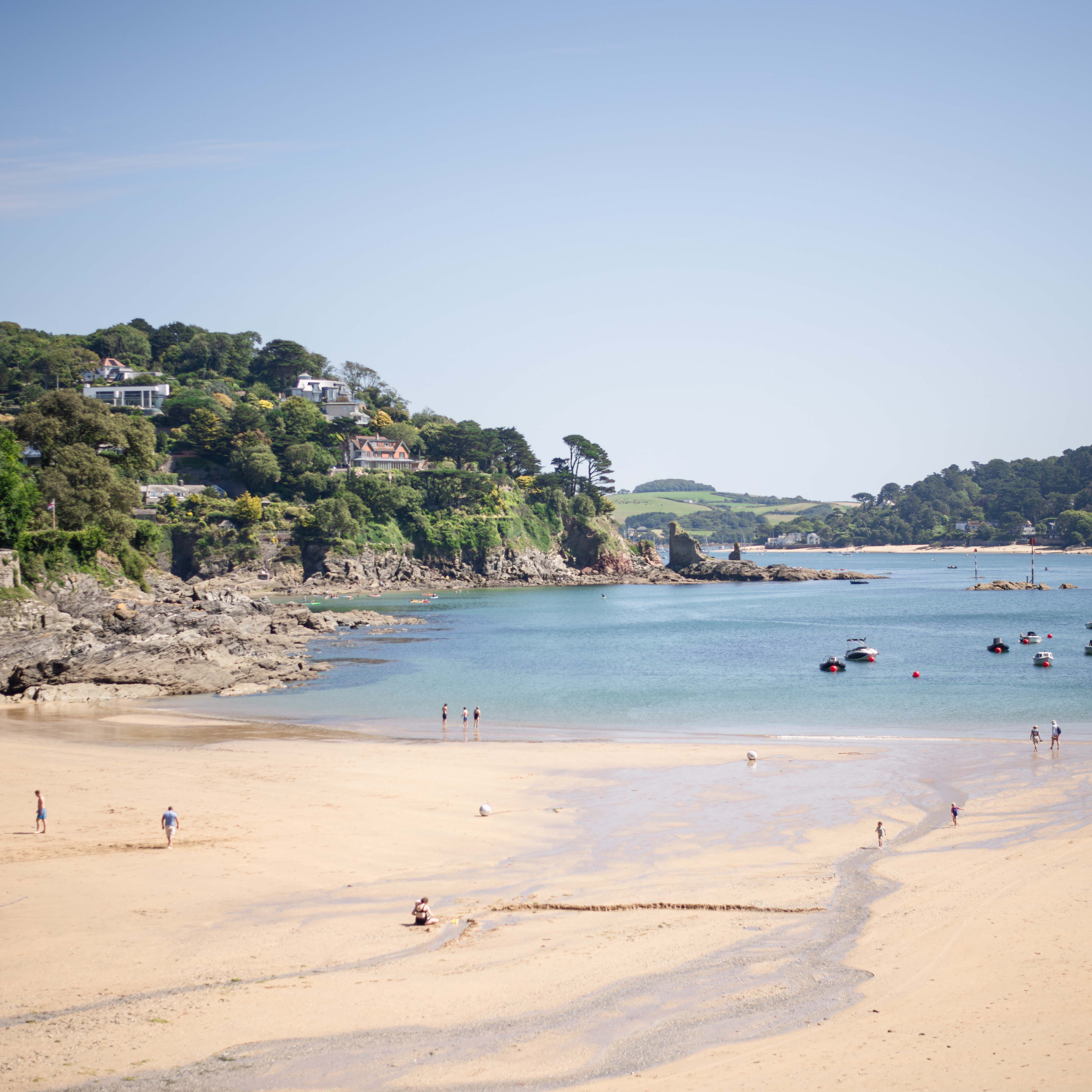 A sandy beach with people walking and relaxing, a calm blue sea with boats, and green hills with houses in the background.