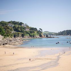 A sandy beach with people walking and relaxing, a calm blue sea with boats, and green hills with houses in the background.