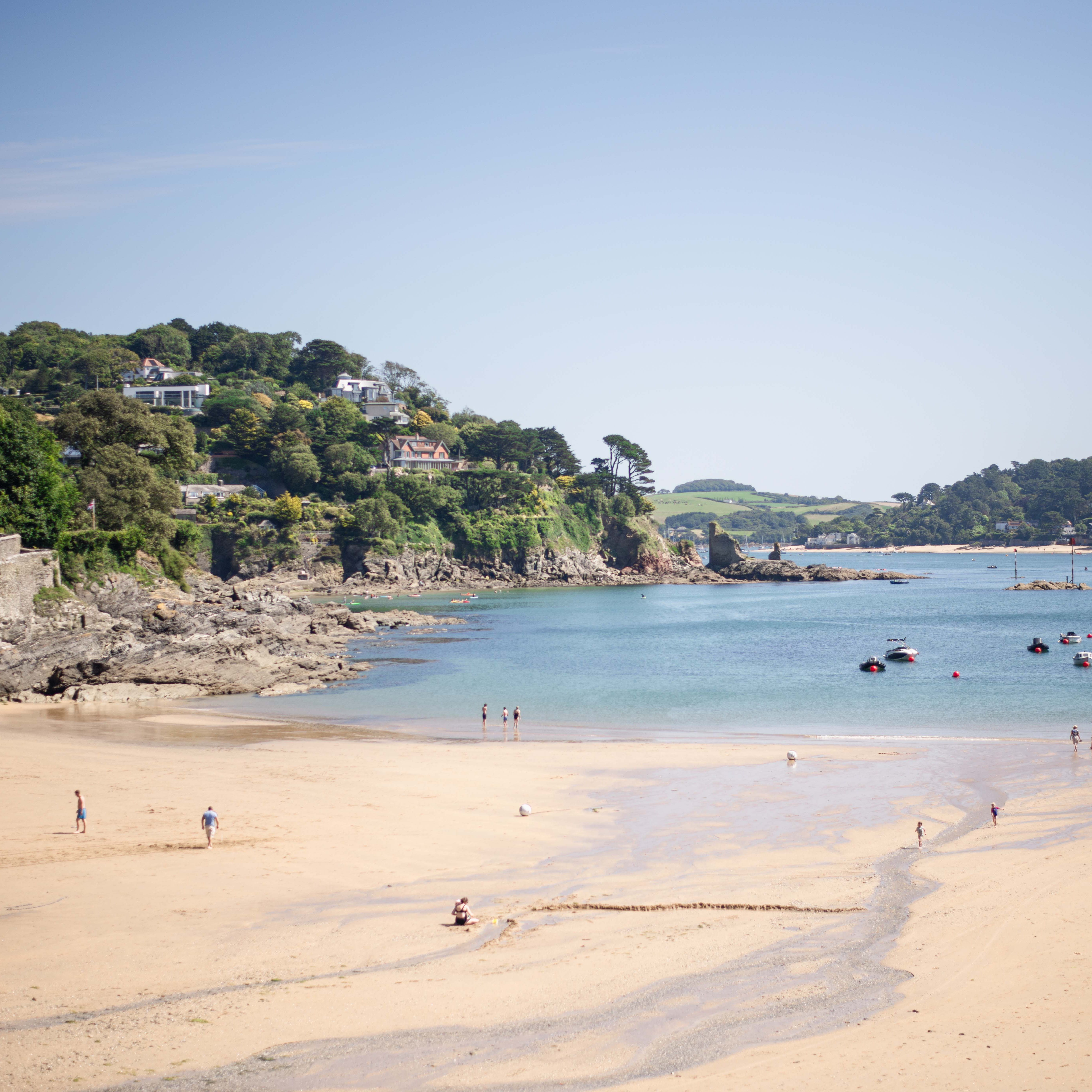 A sandy beach with people walking and relaxing, a calm blue sea with boats, and green hills with houses in the background.