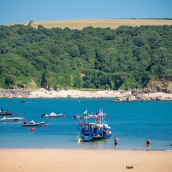 A beach with boats on blue water, people on the sand, and green hills in the background.