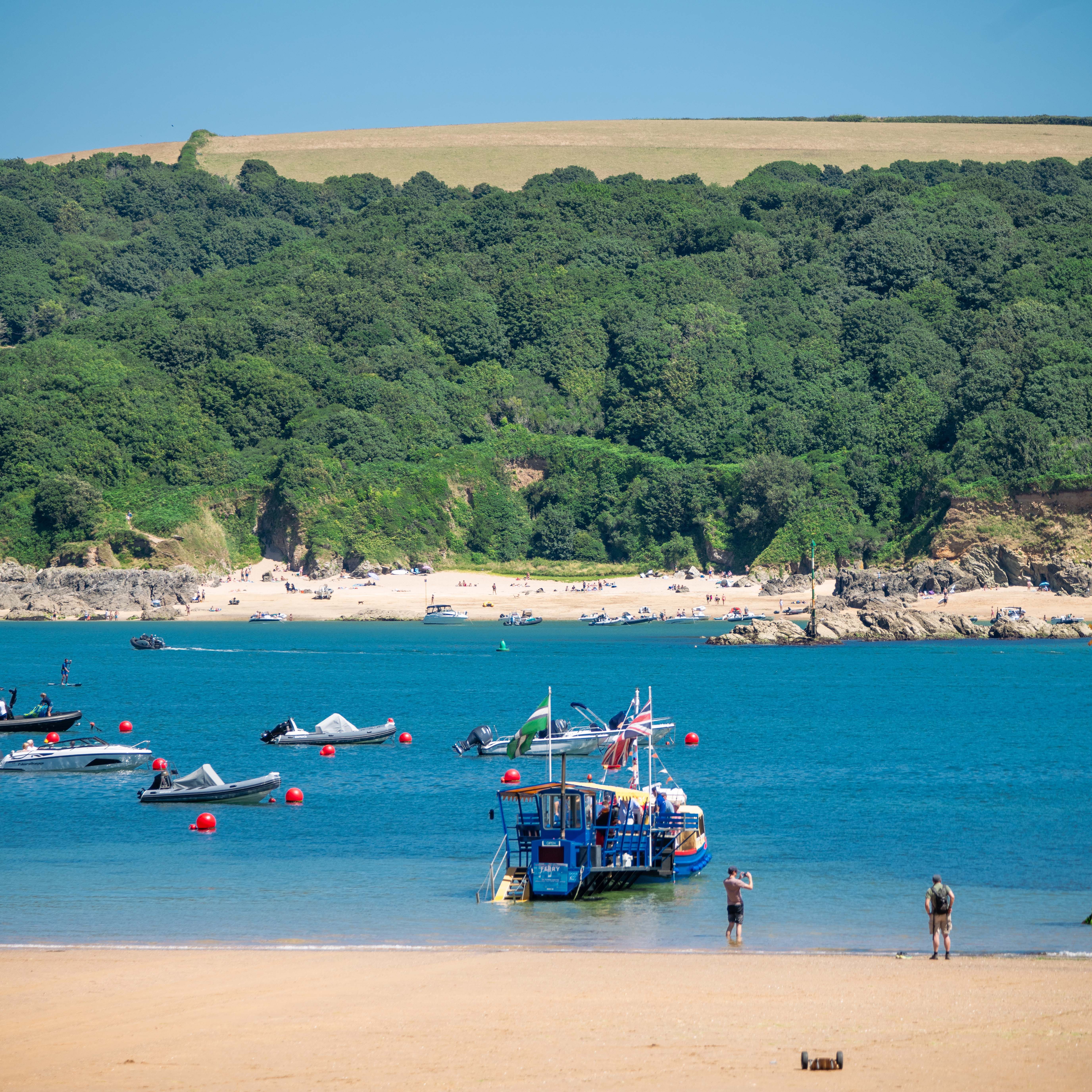 A beach with boats on blue water, people on the sand, and green hills in the background.