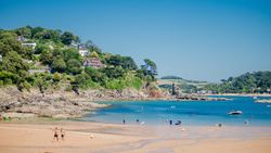 A scenic coastal beach with people enjoying the water, boats, and houses on a lush green hillside.