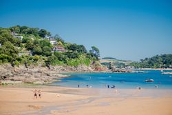 A scenic coastal beach with people enjoying the water, boats, and houses on a lush green hillside.