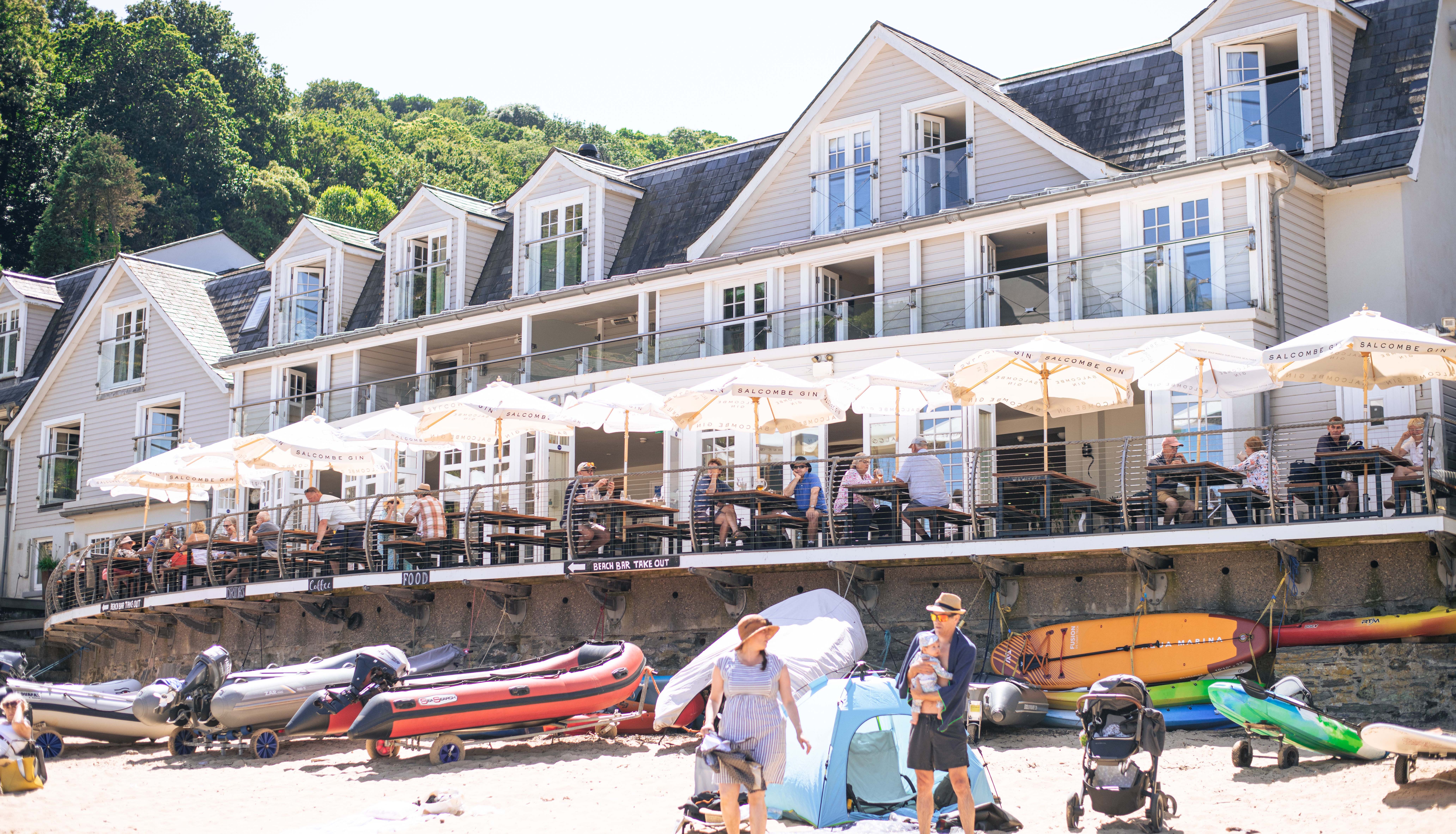 Beachfront hotel with outdoor dining, people relaxing under umbrellas, inflatable boats and kayaks on sandy beach, and a family with a baby and stroller.