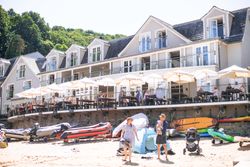 Beachfront hotel with outdoor dining, people relaxing under umbrellas, inflatable boats and kayaks on sandy beach, and a family with a baby and stroller.