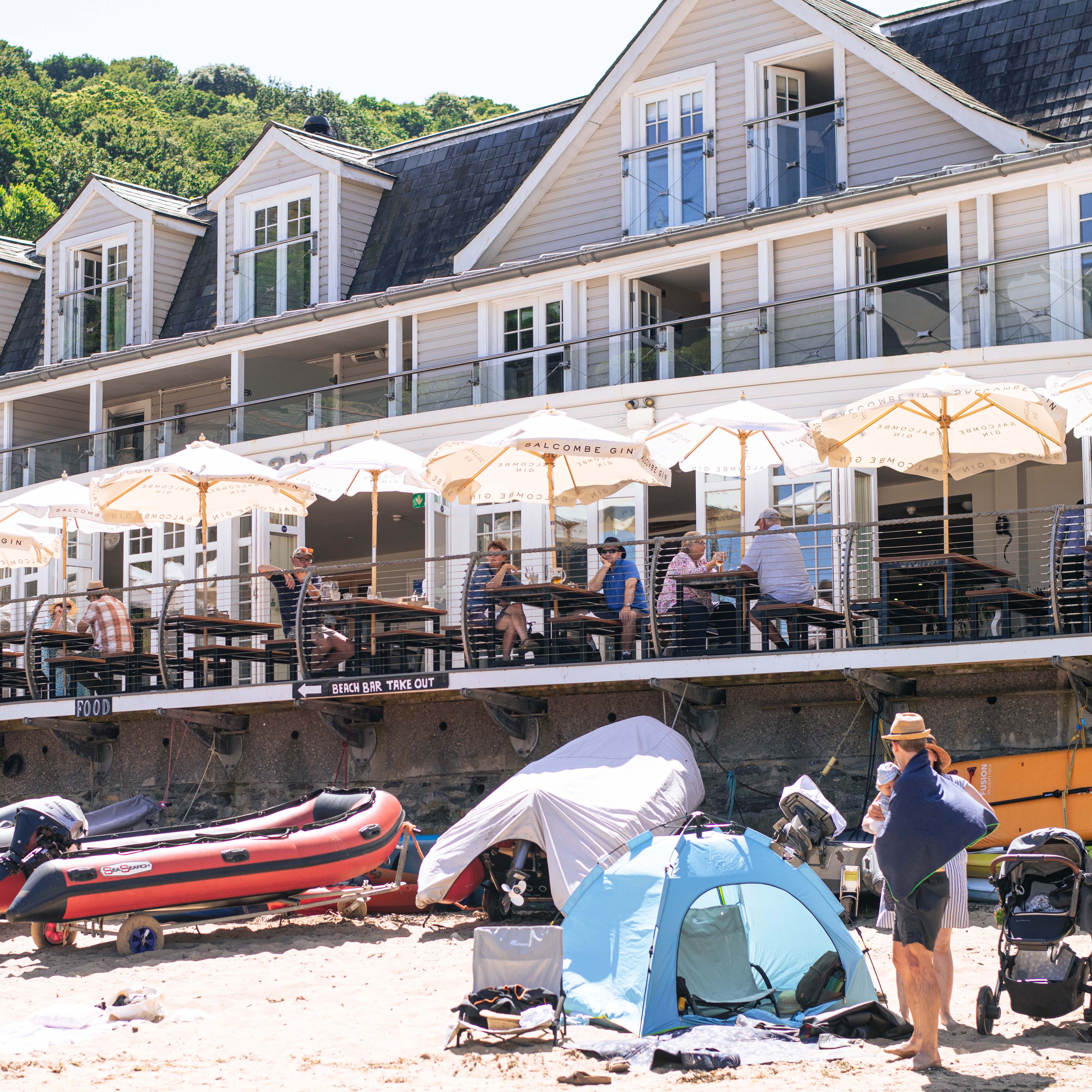 Beachfront restaurant with outdoor seating, umbrellas, and people dining, with inflatable boats and tents on the sandy beach below.