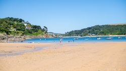 People enjoying a sandy beach with boats floating on blue water and green hills in the background.