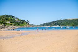 People enjoying a sandy beach with boats floating on blue water and green hills in the background.