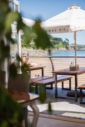 Outdoor café seating area overlooking a beach and water, with wooden tables, a large white umbrella, and some greenery in the foreground.