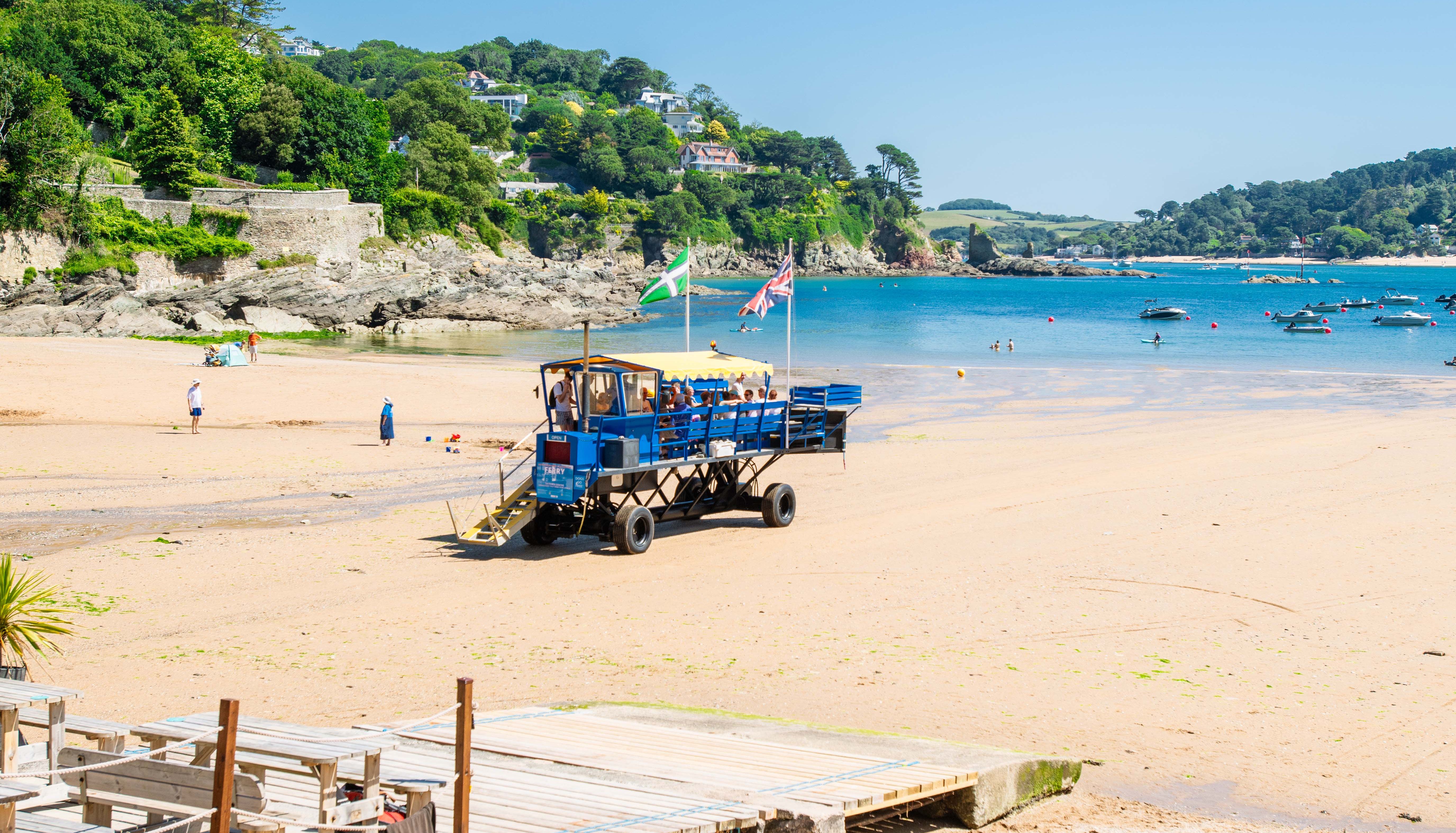 A sandy beach with a blue amphibious vehicle, flags, a wooden walkway, and water sports equipment in the foreground. Green hills and houses line the coast in the background.