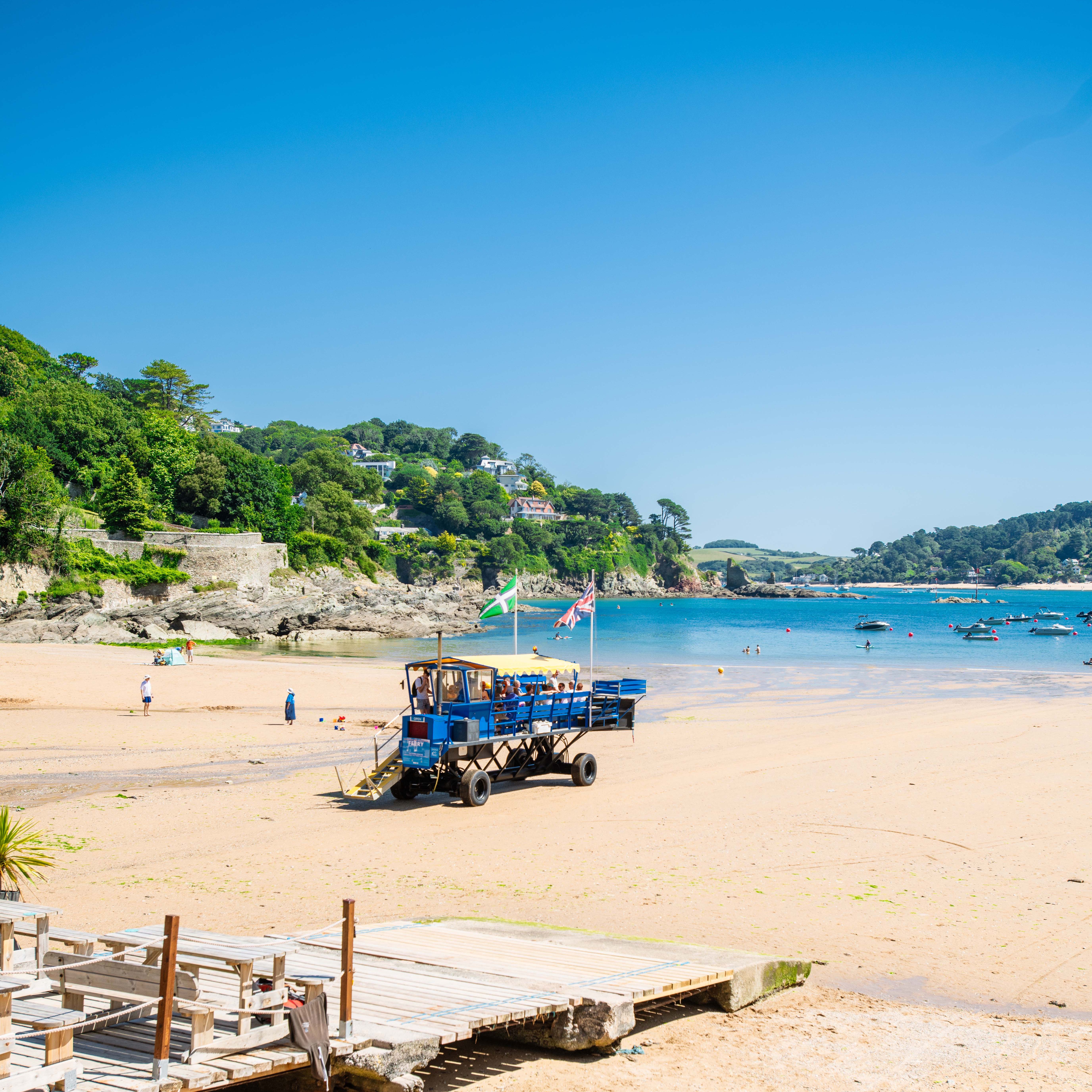 A sandy beach with a blue amphibious vehicle, flags, a wooden walkway, and water sports equipment in the foreground. Green hills and houses line the coast in the background.