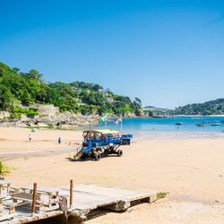 A sandy beach with a blue amphibious vehicle, flags, a wooden walkway, and water sports equipment in the foreground. Green hills and houses line the coast in the background.