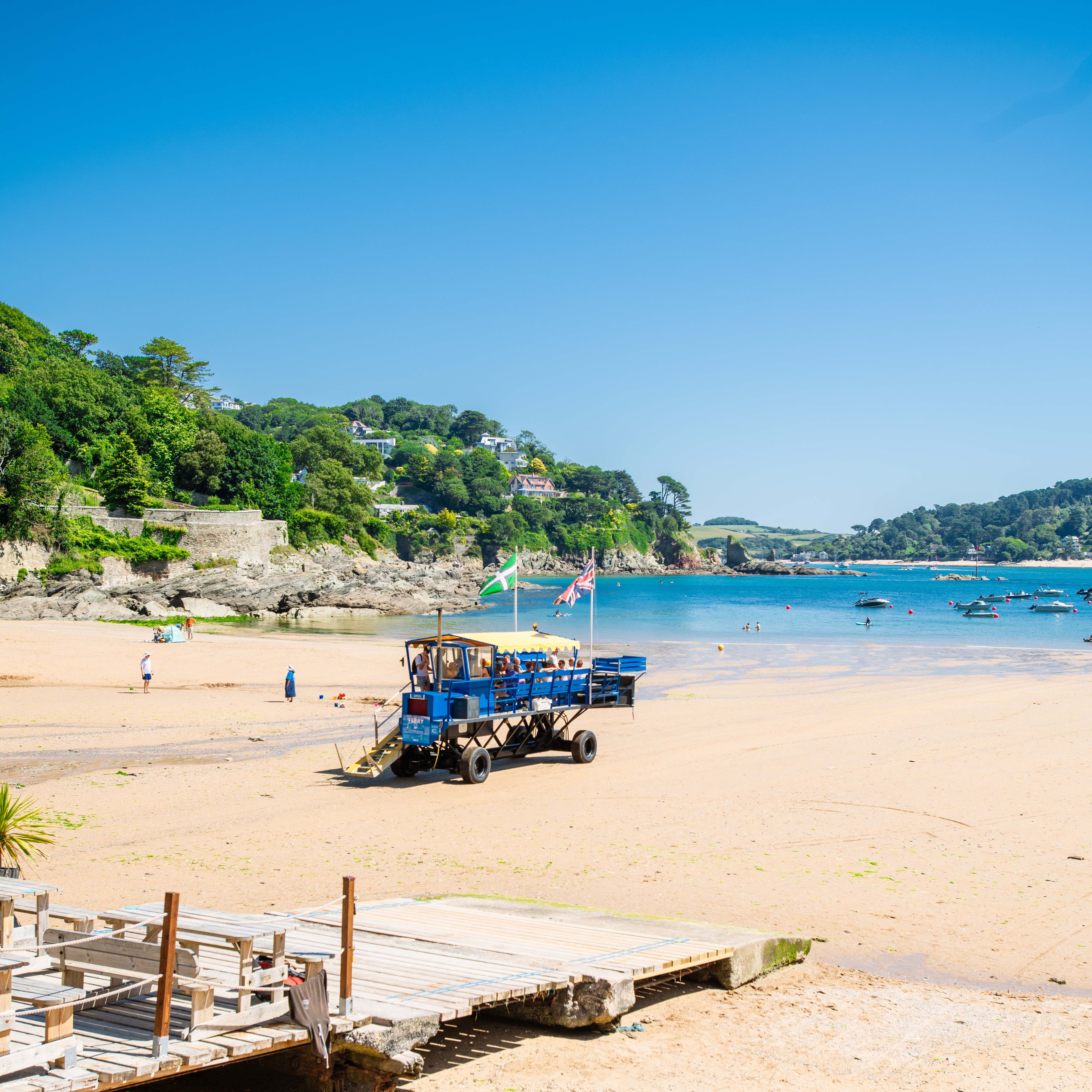 A sandy beach with a blue amphibious vehicle, flags, a wooden walkway, and water sports equipment in the foreground. Green hills and houses line the coast in the background.