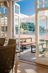 Sunny beachfront patio viewed through open glass doors of a restaurant, with wooden picnic tables, large white umbrellas, and a scenic coastline in the background.