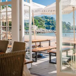 Sunny beachfront patio viewed through open glass doors of a restaurant, with wooden picnic tables, large white umbrellas, and a scenic coastline in the background.