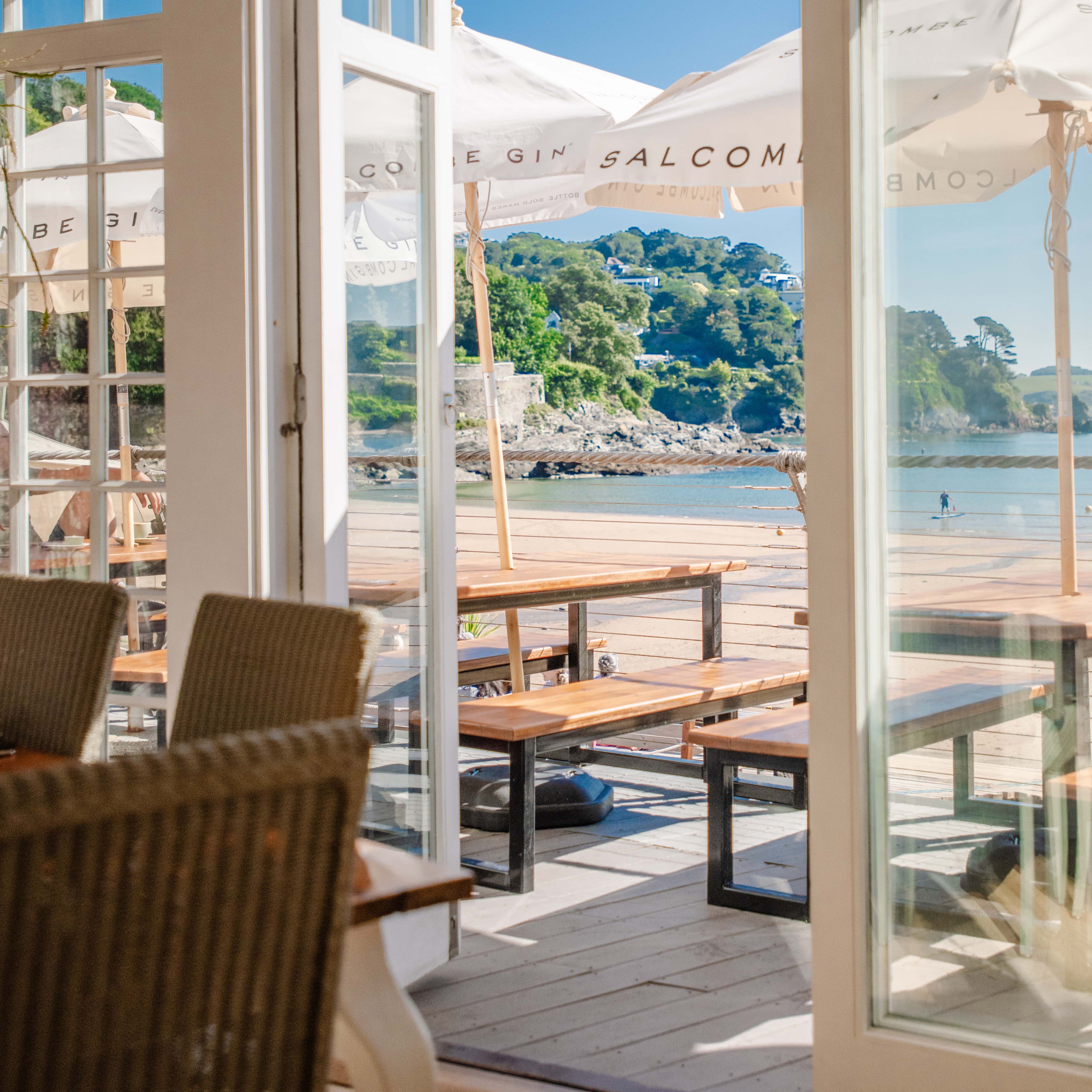 Sunny beachfront patio viewed through open glass doors of a restaurant, with wooden picnic tables, large white umbrellas, and a scenic coastline in the background.