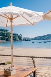 Beachfront café table under an umbrella with a view of the sandy beach, calm blue water, boats, and green hills in the background.