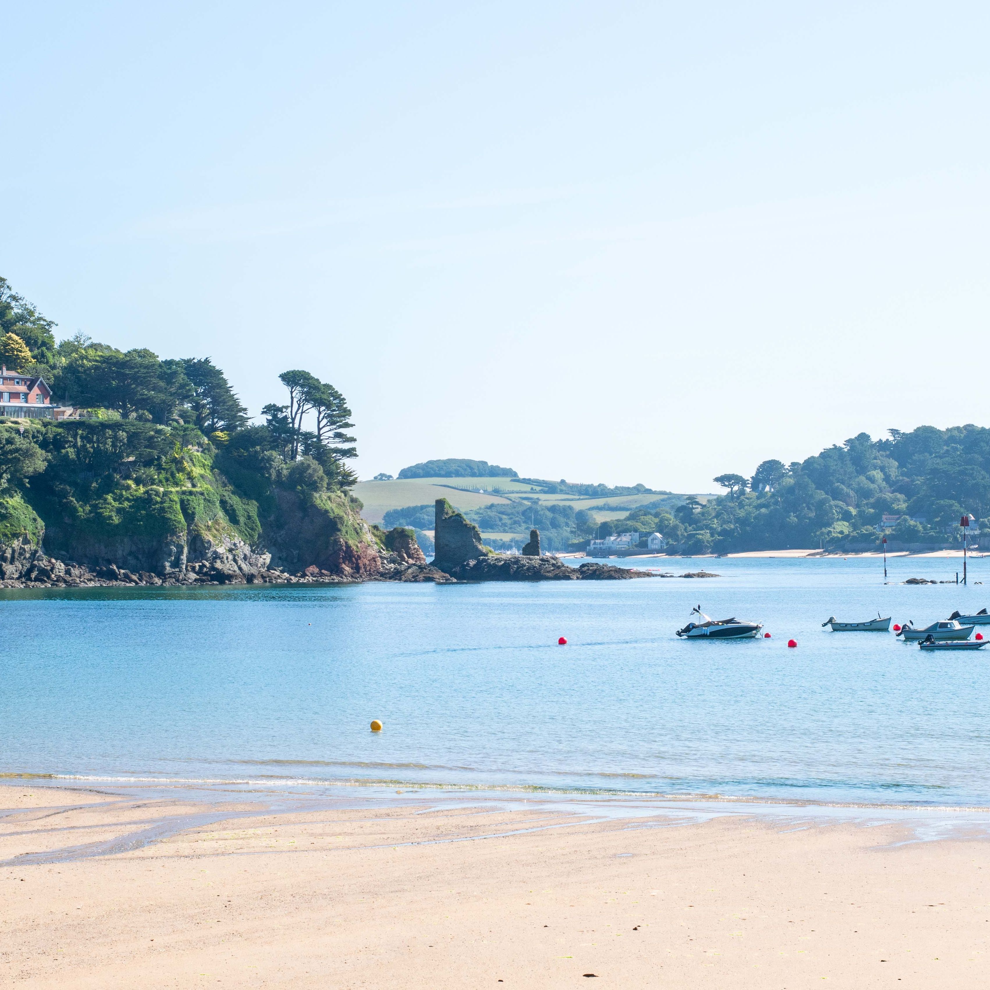 Sunny beach with clear blue water, boats anchored offshore, and green hills in the background