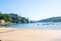 Sunny beach with clear blue water, boats anchored offshore, and green hills in the background