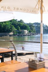 Two children standing on a dock by a calm bay with green hills and stone walls in the background, viewed from a shaded outdoor table area.