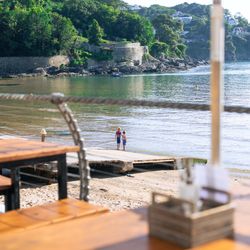 Two children standing on a dock by a calm bay with green hills and stone walls in the background, viewed from a shaded outdoor table area.