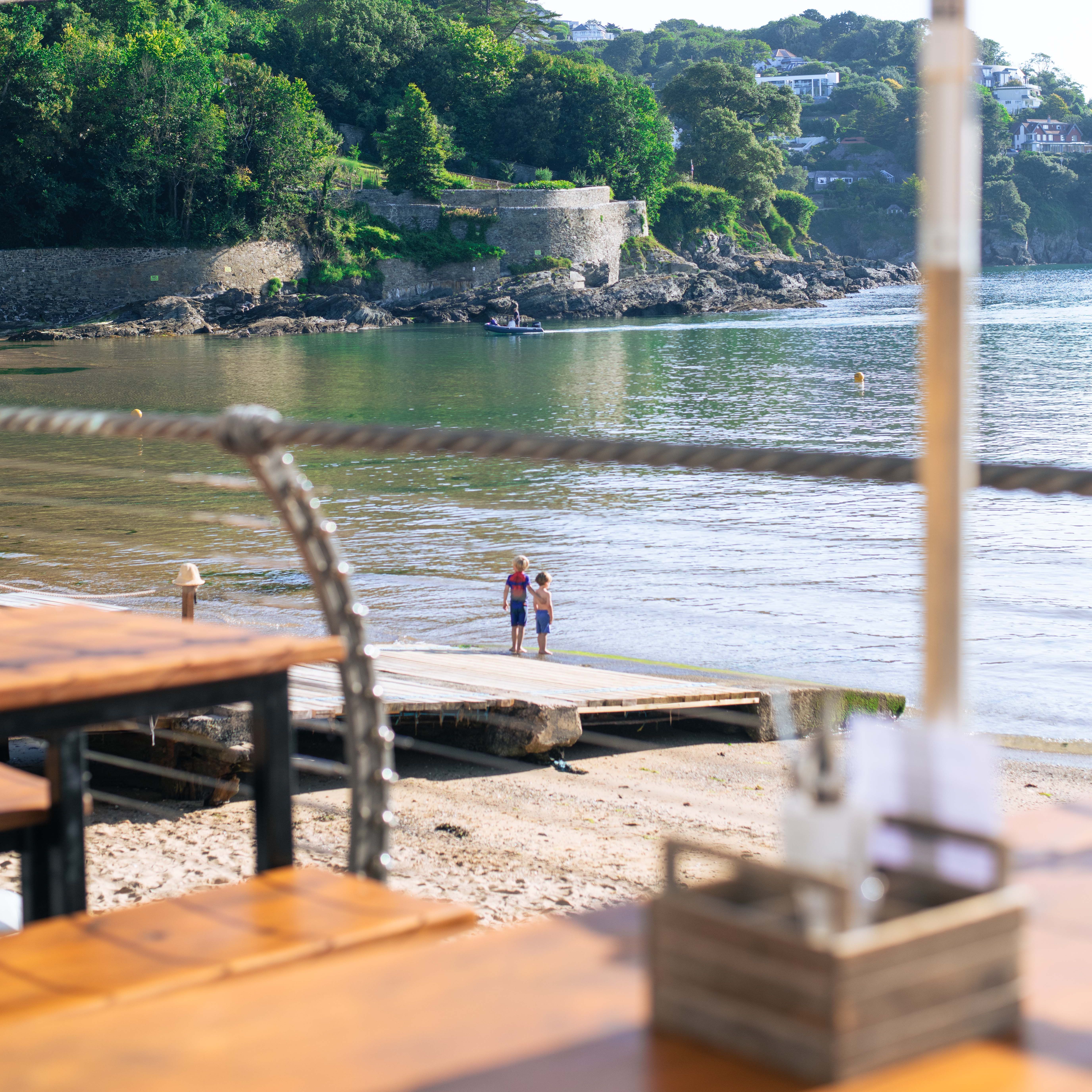 Two children standing on a dock by a calm bay with green hills and stone walls in the background, viewed from a shaded outdoor table area.