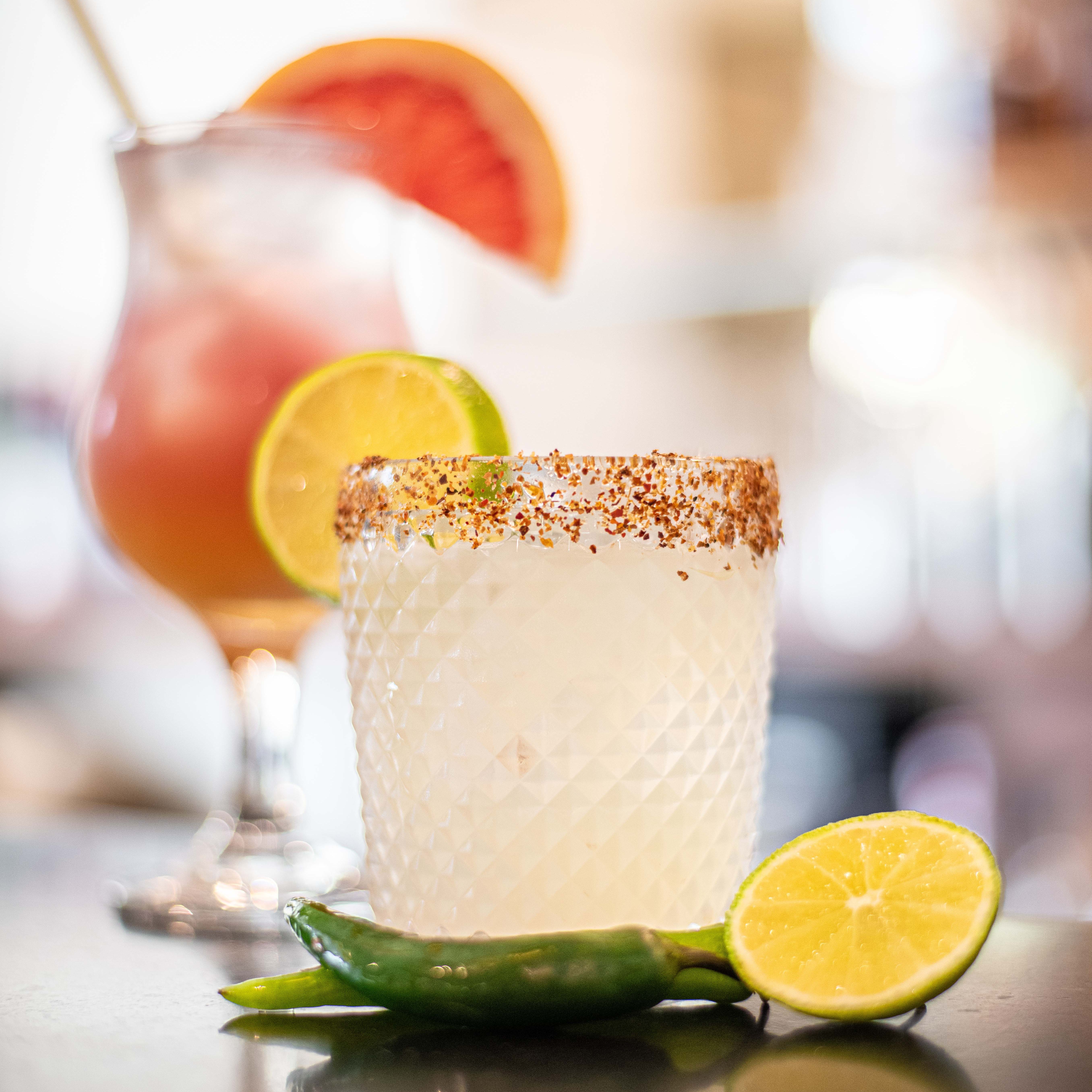 A close-up of a cocktail with a chili pepper, lime, and spicy rim, with another cocktail and garnish in the blurred background.