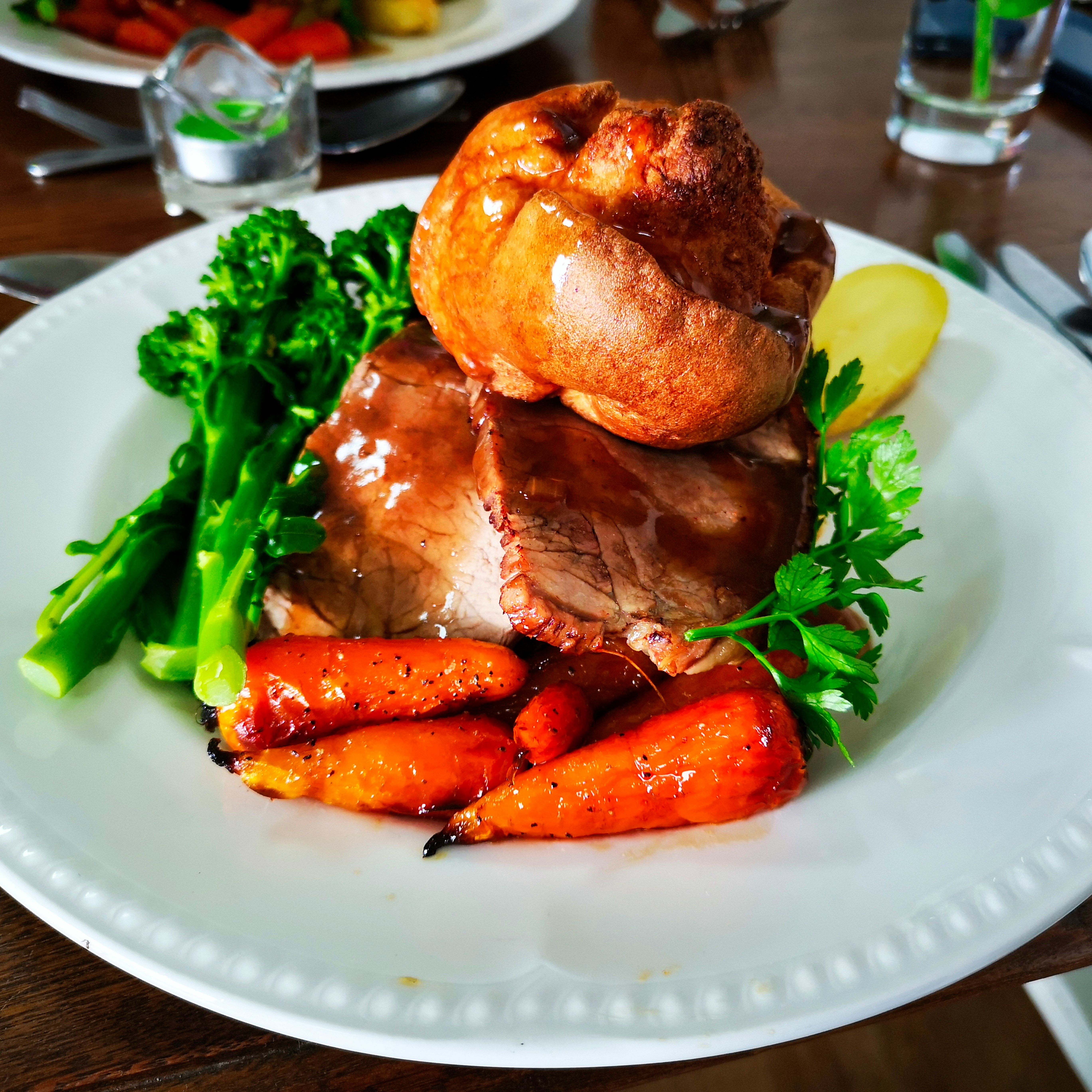 Traditional British roast dinner with roast beef, Yorkshire pudding, carrots, broccoli, and parsley on a white plate.