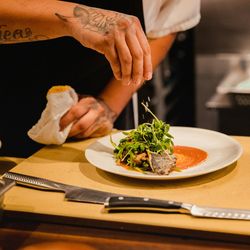 Chef garnishing a plated dish of greens and fish with sauce