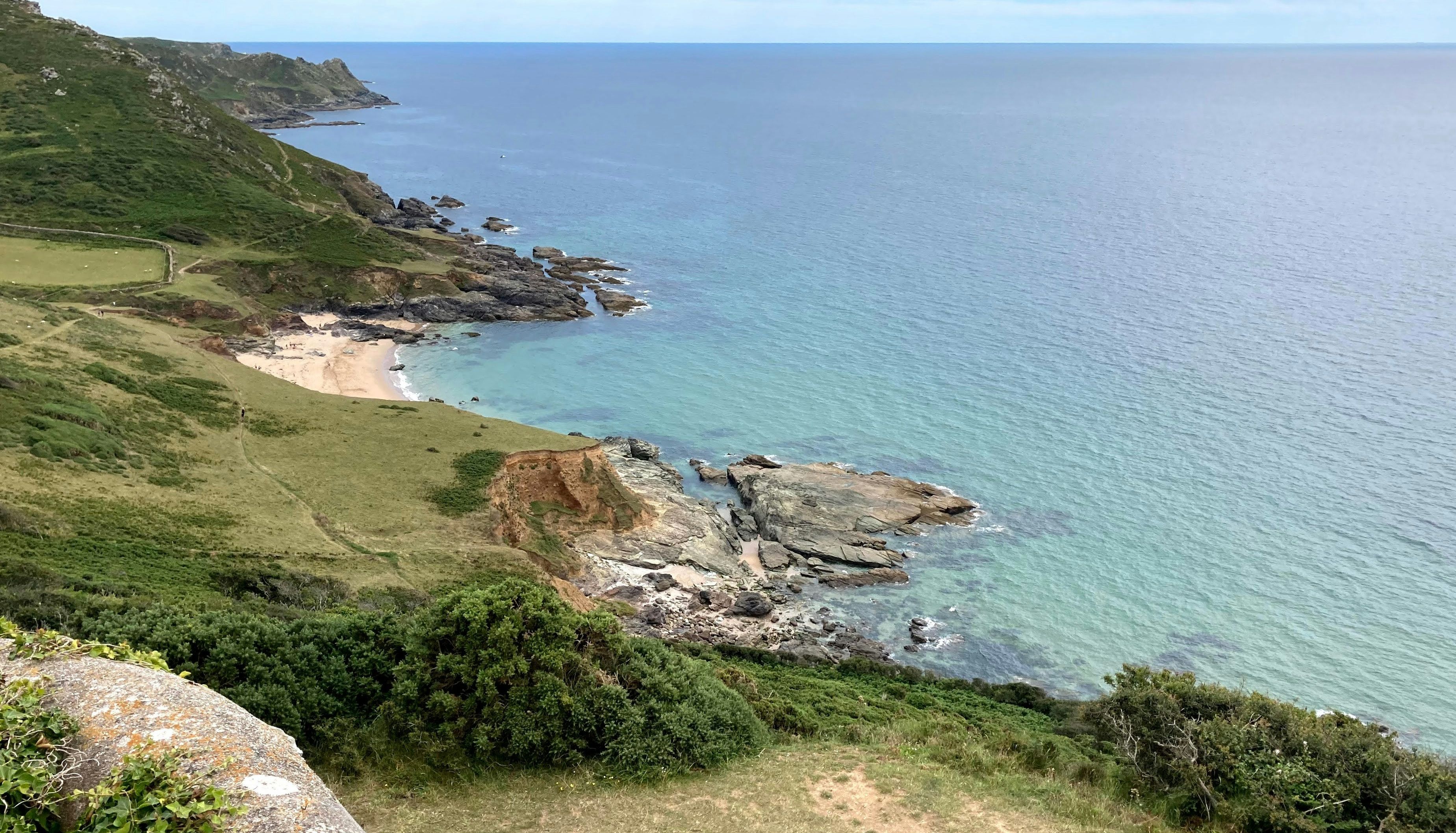A scenic coastal view showing green cliffs, rocky shores, and a sandy beach meeting clear blue ocean water under a partly cloudy sky.