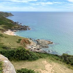 A scenic coastal view showing green cliffs, rocky shores, and a sandy beach meeting clear blue ocean water under a partly cloudy sky.