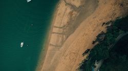 Aerial view of a sandy beach with boats on water and vehicles parked near the shoreline